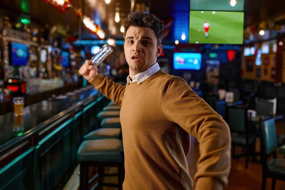 Ein Mann in einem braunen Pullover hebt ein Glas in einer belebten Bar mit Fernsehern. Die Szene wirkt angespannt.