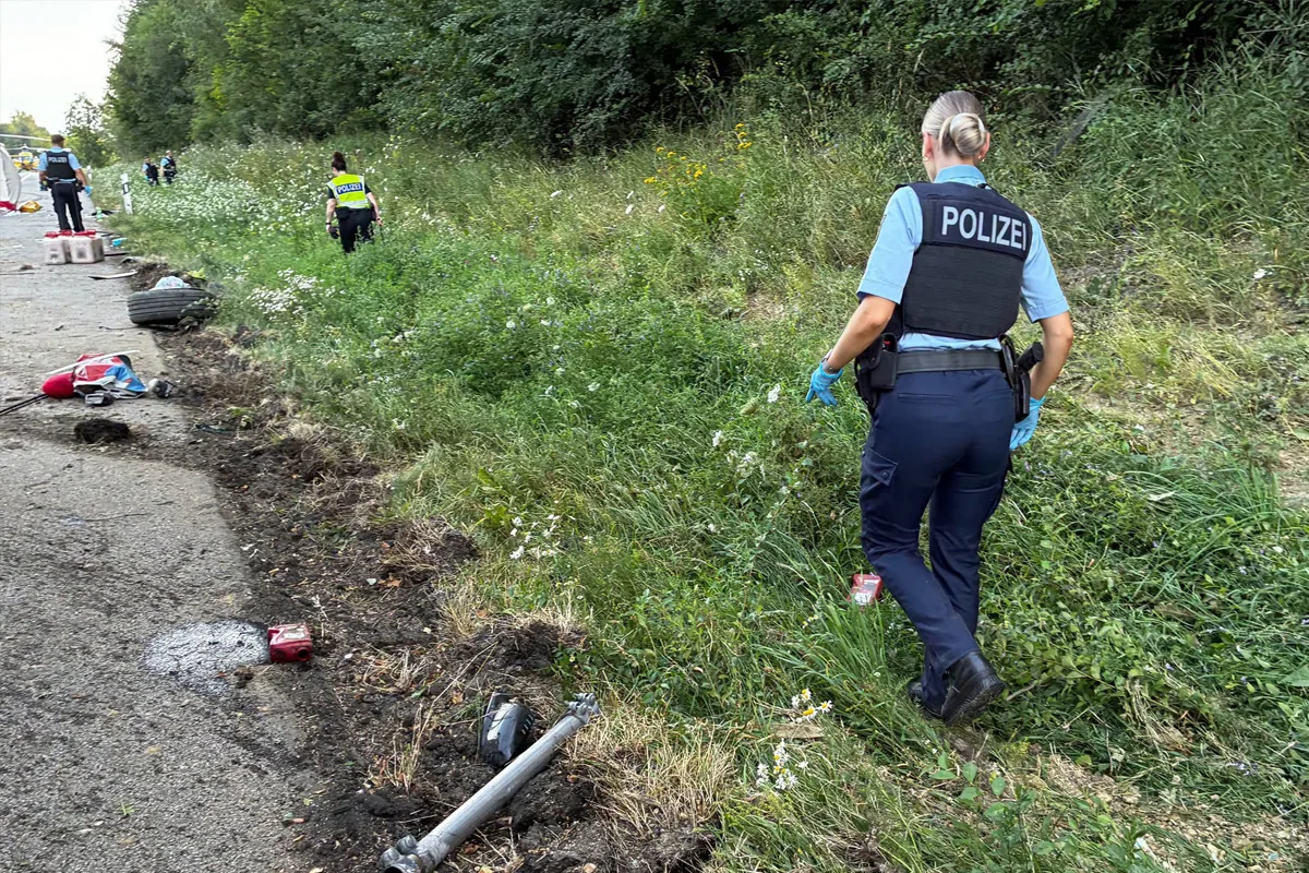 Mehrere Polizisten, darunter eine Frau im Vordergrund, durchsuchen das grasbewachsene Gebiet neben einer Straße. Abgelegte Objekte und beschädigte Fahrzeugteile sind sichtbar.