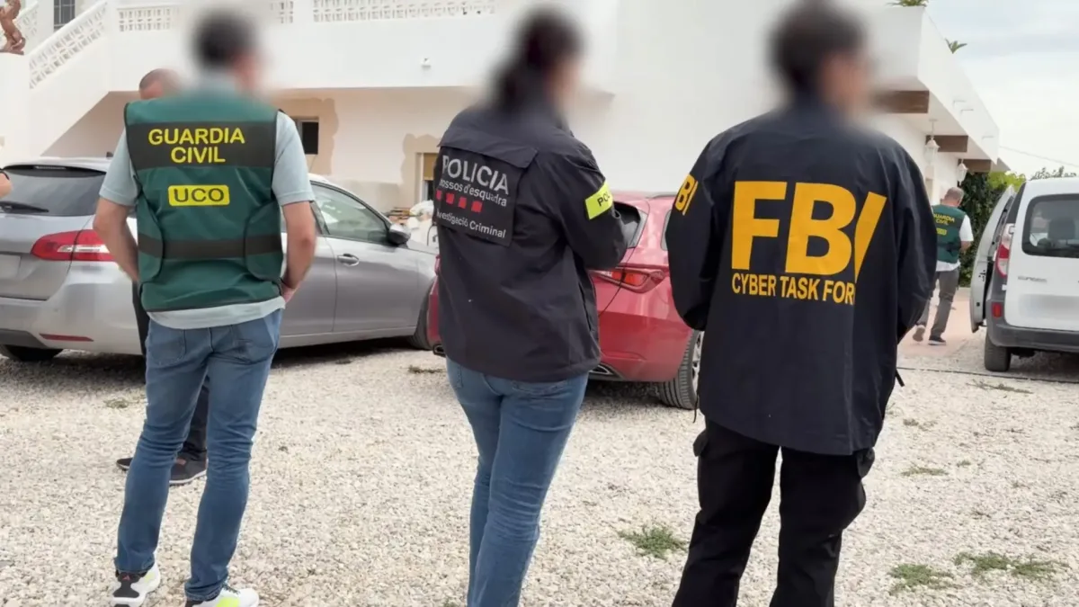 Drei Personen in Jacken der Guardia Civil, Policia und FBI stehen in einer Hofeinfahrt neben geparkten Autos.