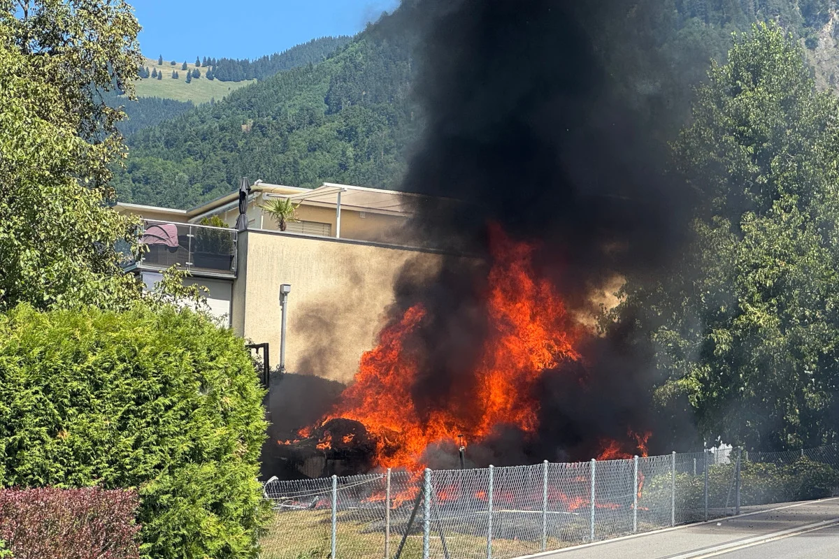 Ein großes, intensives Feuer lodert hinter einem Wohngebäude, dichte Rauchschwaden steigen in den Himmel. Im Vordergrund ist ein Zaun sichtbar.