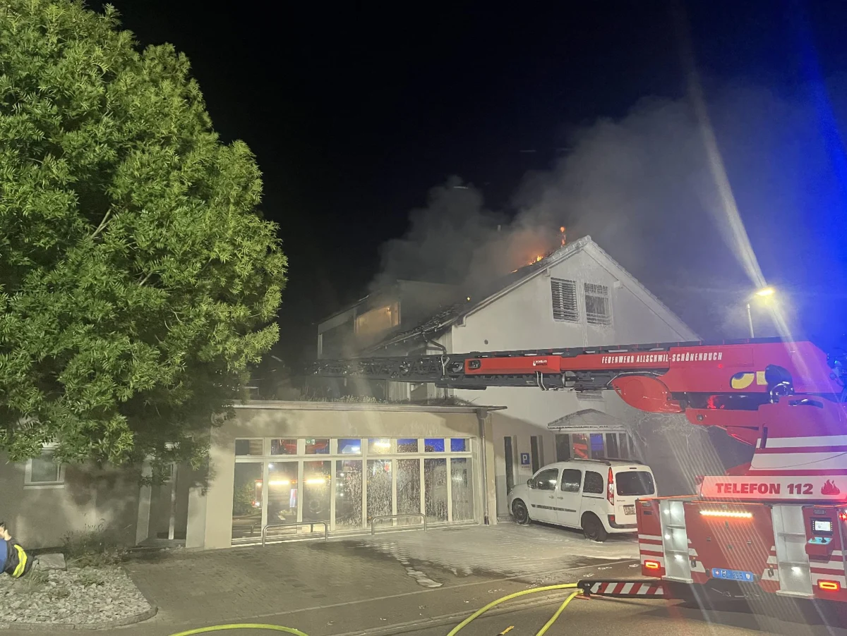 Ein Wohnhaus steht bei Nacht in Flammen, während ein Feuerwehrfahrzeug mit Leiter im Vordergrund den Brand bekämpft. Dicker Rauch steigt vom Dach auf und Lichter strahlen hell.