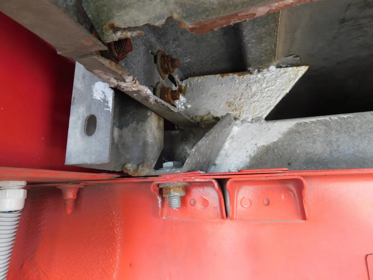 Das Bild zeigt eine Nahaufnahme von rostigen Schrauben und einem Metallträger nahe einer roten Lackierung. Der Rost ist stark ausgeprägt und deutet auf Korrosion hin.