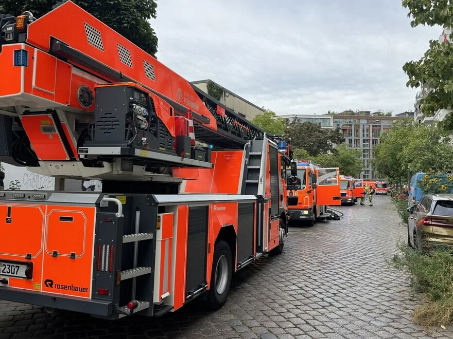 Mehrere orangefarbene Feuerwehrfahrzeuge sind auf einer gepflasterten Stadtstraße geparkt. Ein Fahrzeug ist prominent im Vordergrund sichtbar.