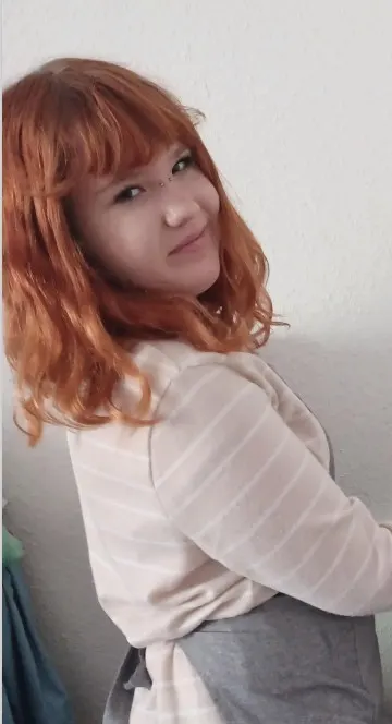 Eine Frau mit roten Haaren und hellem Oberteil schaut über die Schulter in Richtung Kamera. Der Hintergrund ist weiß und schlicht.