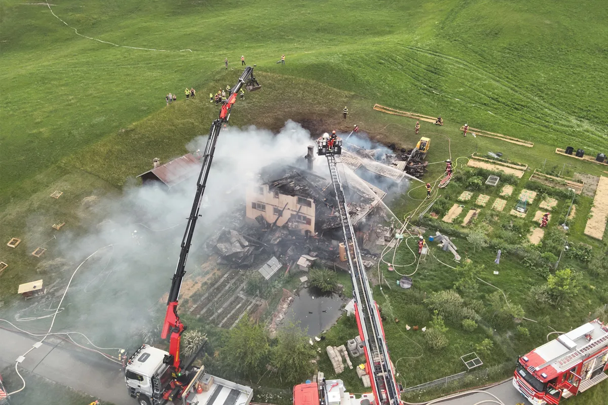 Eine Luftaufnahme zeigt ein brennendes Haus, das von Feuerwehrfahrzeugen und Einsatzkräften umgeben ist. Rauch steigt auf, während Kräne Wasser auf das Gebäude sprühen.