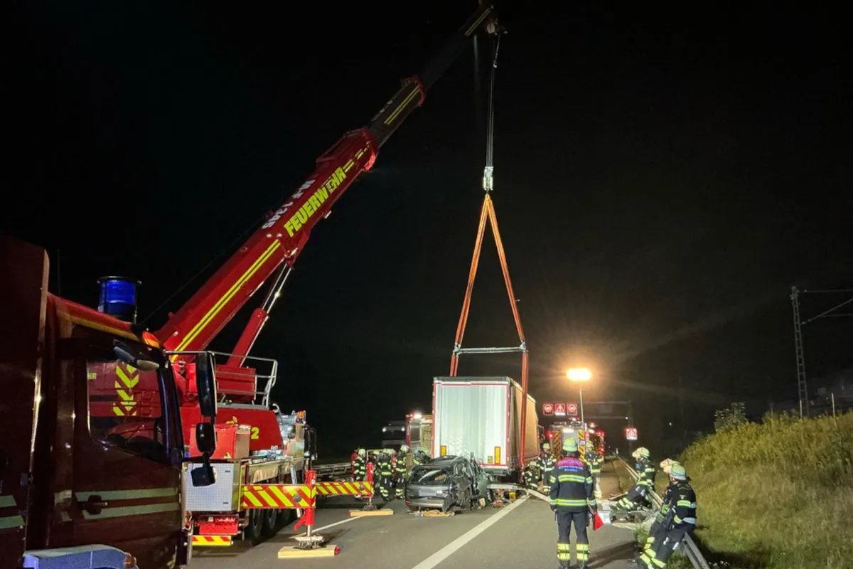 Ein großer roter Kran hebt einen Lkw-Anhänger auf einer Autobahn an. Mehrere Feuerwehrleute stehen daneben und beobachten den Vorgang bei Nacht.