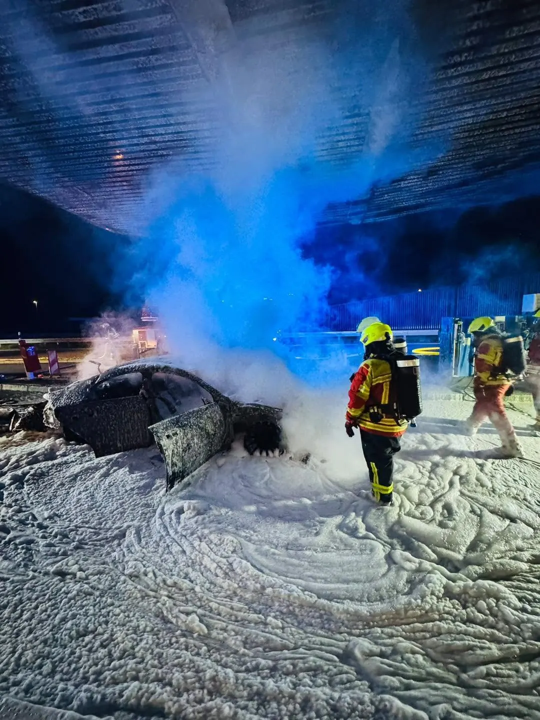 Firefighters in protective gear stand amidst thick firefighting foam, extinguishing a car fire under a bridge at night.