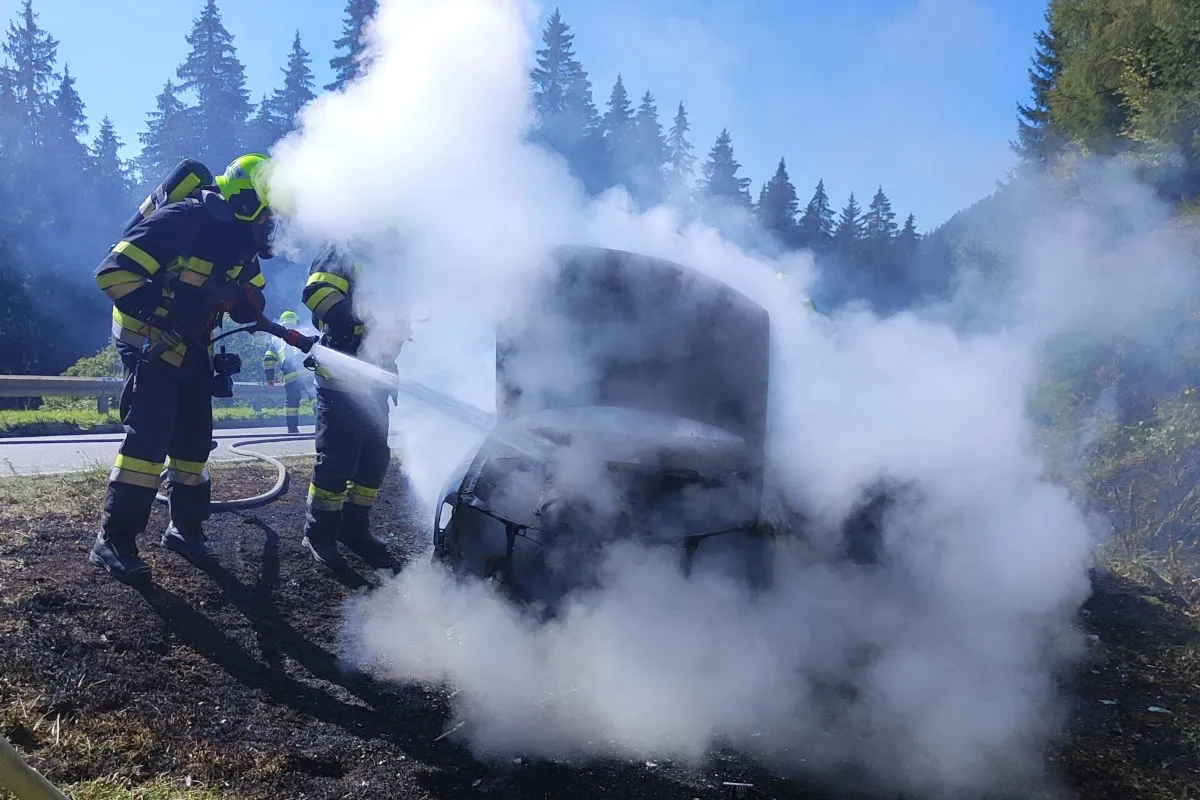 Zwei Feuerwehrleute in Schutzanzügen bekämpfen einen Autoverbrand mit Wasserschläuchen auf einer landschaftlichen Straße. Dichter Rauch ist um das Fahrzeug sichtbar.