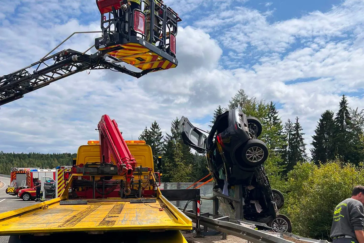 Ein verunfalltes Auto wird von einem Abschleppwagen und Feuerwehrfahrzeugen geborgen. Die Szene spielt sich auf einer Landstraße ab.
