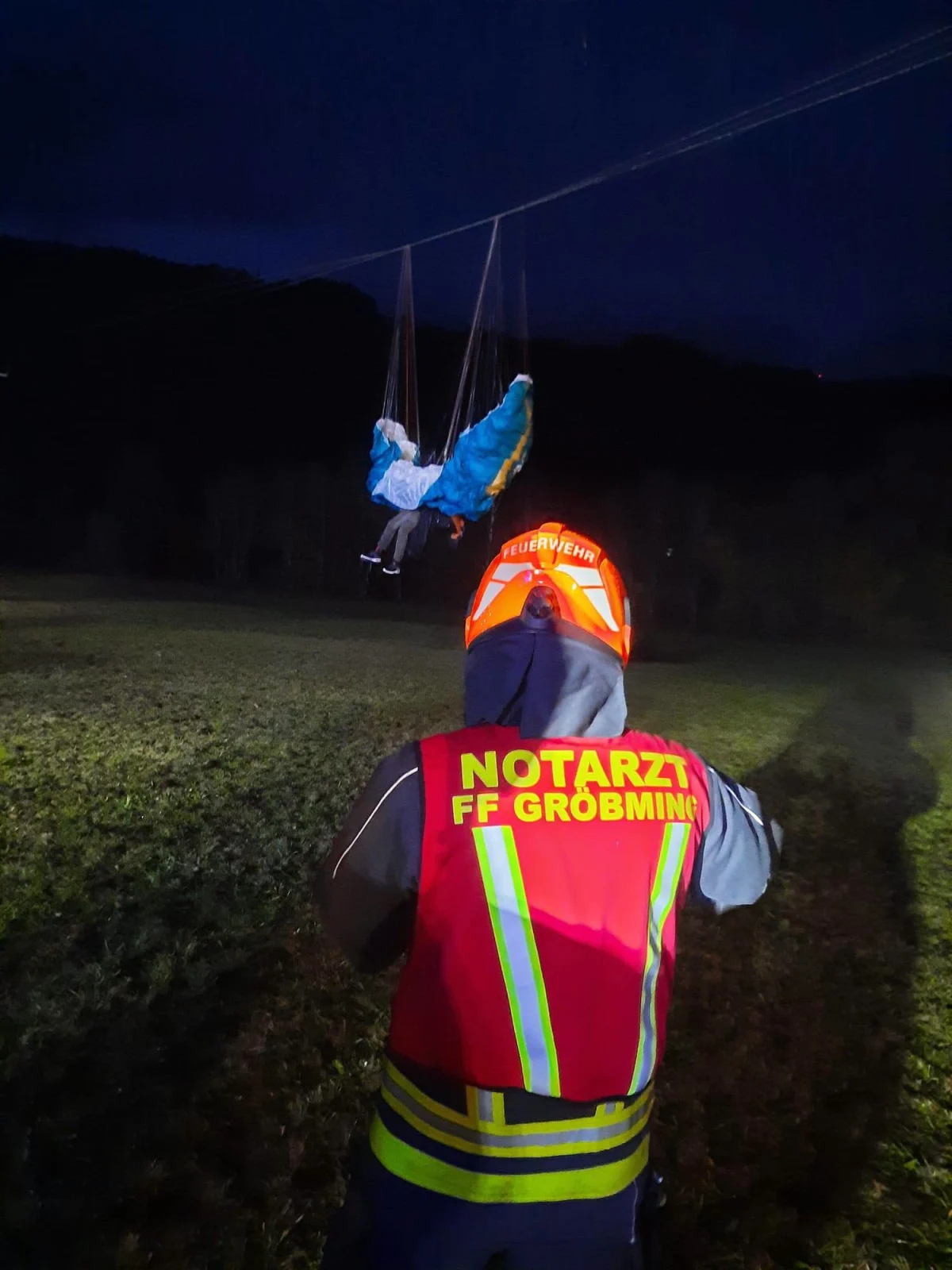 Ein Paragleiter hängt bei Nacht in Stromleitungen fest, während ein Notarzt mit Schutzweste im Vordergrund zu sehen ist.