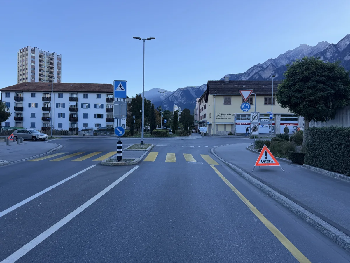Ein Unfallschild steht am Kreisverkehr; im Hintergrund sind Berge und Wohngebäude zu sehen, die auf einen Stadtrand hindeuten.
