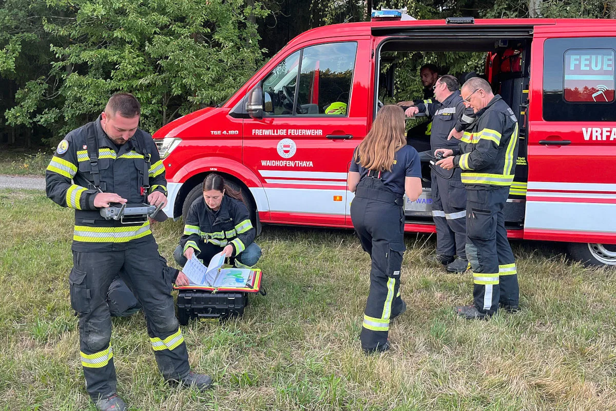 Eine Gruppe von Feuerwehrleuten in Schutzkleidung ist auf einer Wiese bei einem Einsatz neben einem roten Einsatzfahrzeug der Feuerwehr.