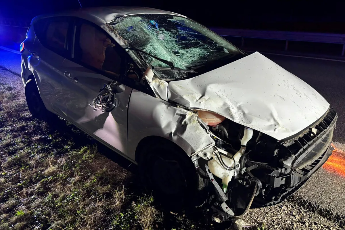 Ein stark beschädigtes weißes Auto mit einer zersplitterten Windschutzscheibe steht am Rand einer Autobahn bei Nachtbeleuchtung. Das Fahrzeug hat erhebliche Beulen und Schäden an der Front und der Seite.