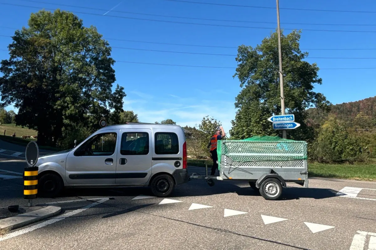 Ein silberner Pkw mit Anhänger steht auf einer Straße nahe eines Wegweisers Richtung Breitenbach und Nunningen. Ein Mann steht neben dem Anhänger.