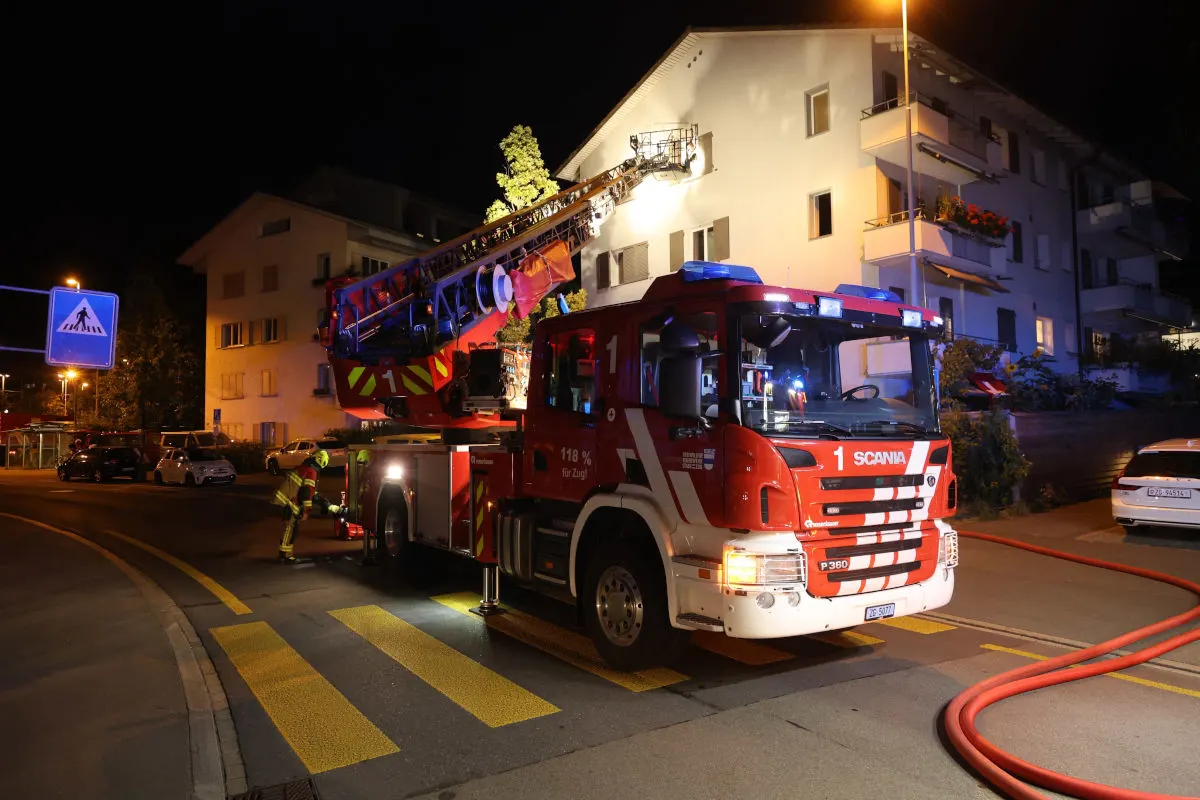 Ein rotes Feuerwehrfahrzeug ist in der Nacht vor einem beleuchteten Wohnhaus stationiert, mit einer Leiter ausgestreckt zur Fassade.