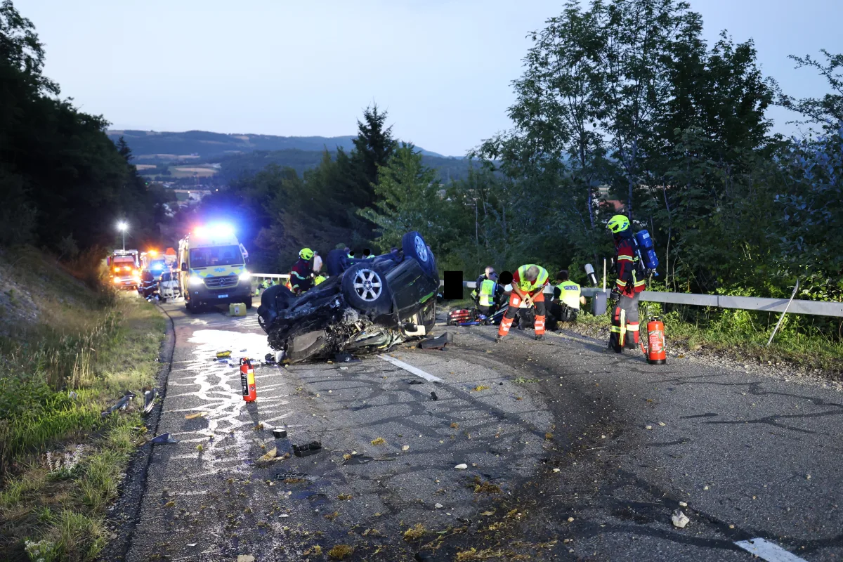 Ein Auto liegt umgestürzt auf einer Landstraße, während mehrere Einsatzkräfte mit Rettungsarbeiten beschäftigt sind. Polizeifahrzeuge stehen mit eingeschaltetem Blaulicht bereit.