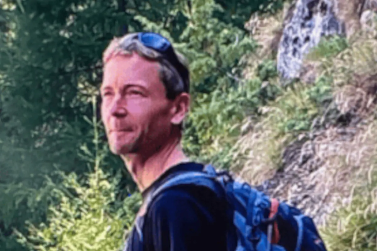 Ein Mann mit Sonnenbrille auf dem Kopf und einem Rucksack wandert auf einem bewachsenen Waldweg bei Tageslicht.