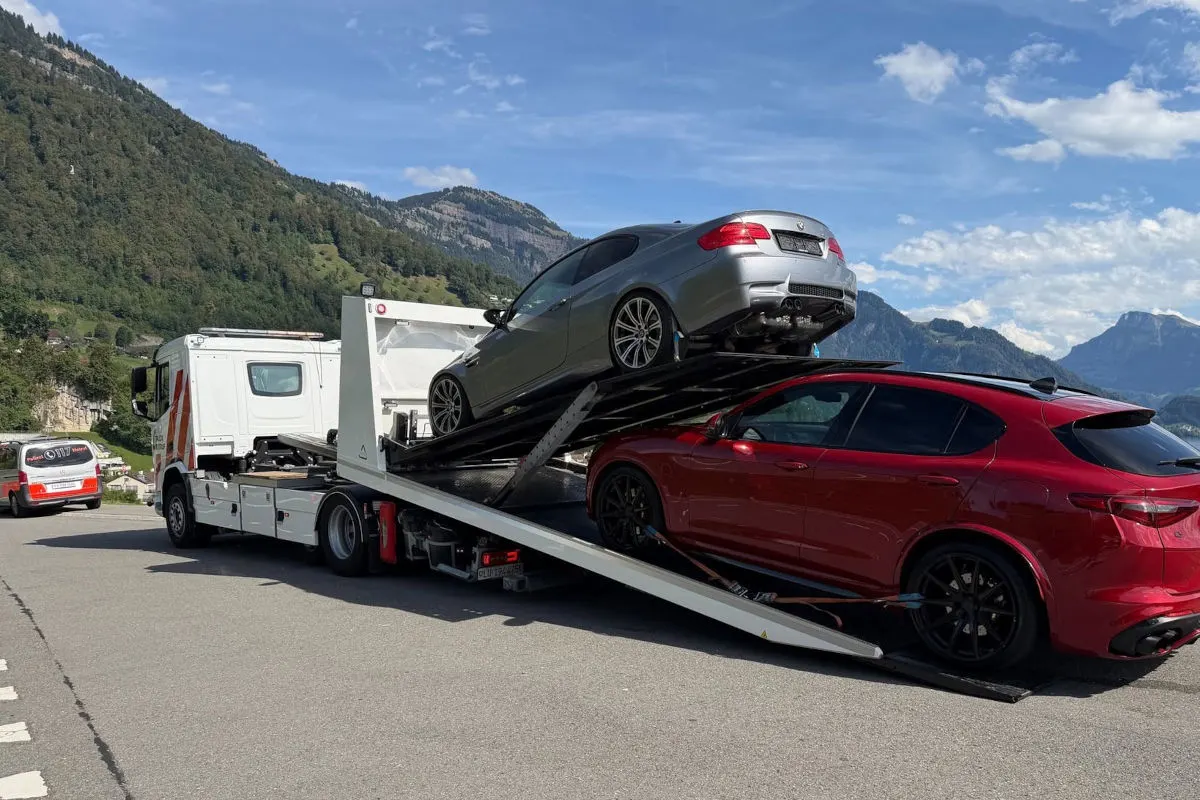 Ein Abschleppwagen transportiert einen silbernen und einen roten Pkw auf einer Straße mit Bergen im Hintergrund. Ein weiteres Fahrzeug ist auf der Straße zu sehen.