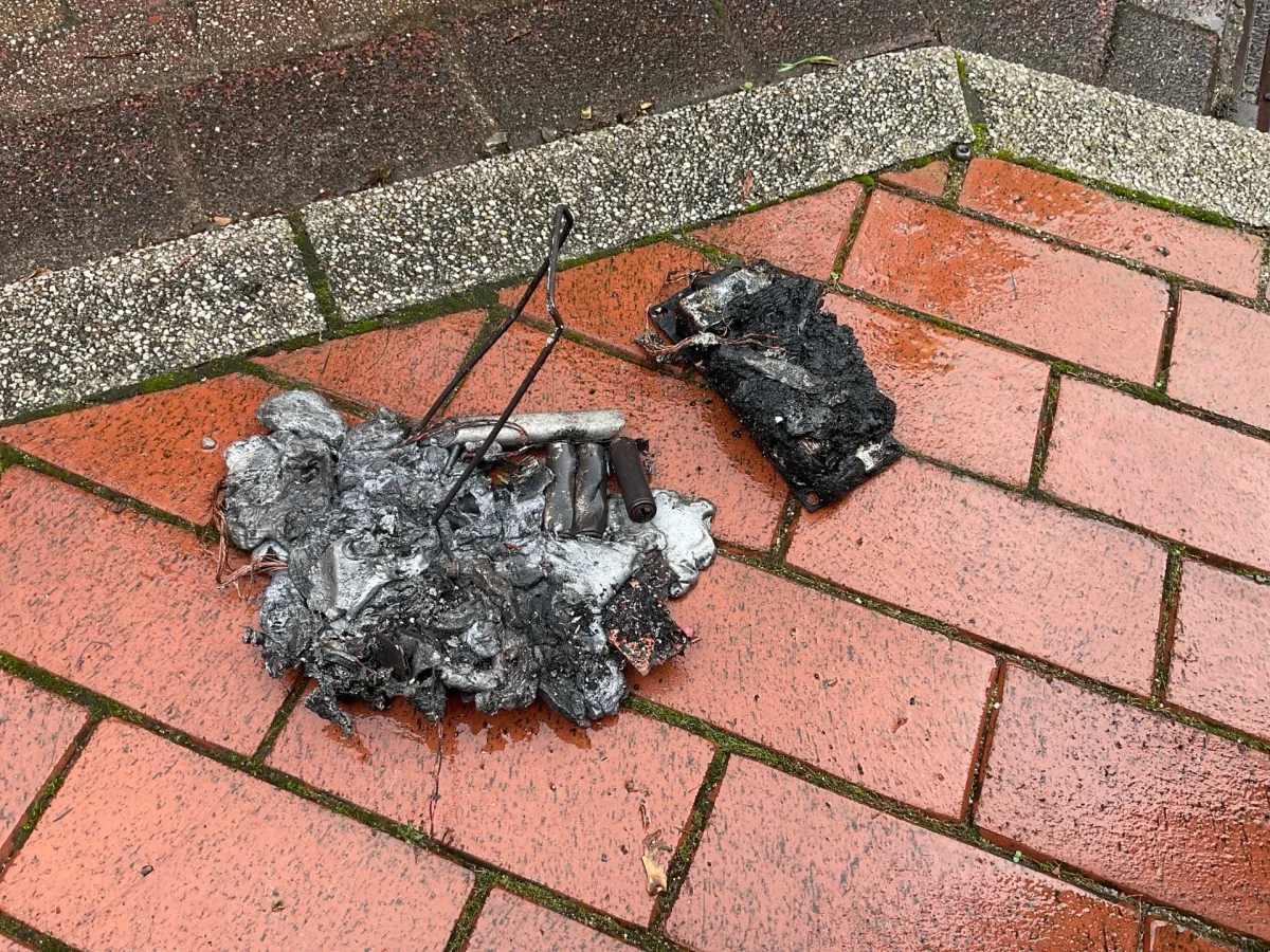 Auf dem Bild sind die verkohlten Überreste eines Geräts auf einem regennassen Gehweg mit roten Fliesen zu sehen. Das Gerät ist kaum zu erkennen und stark beschädigt.