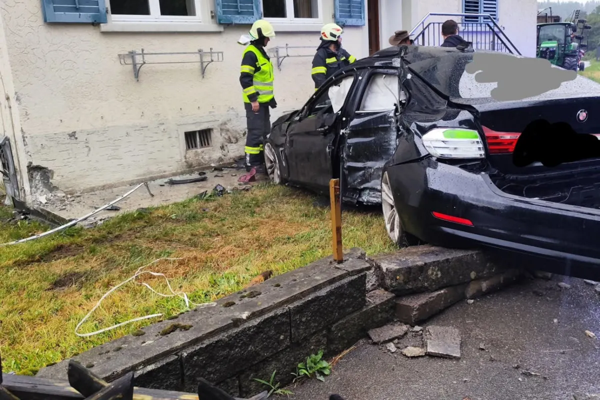 Ein stark beschädigtes schwarzes Auto ist in die Wand eines Wohnhauses geprallt. Feuerwehrleute sind vor Ort, um den Vorfall zu sichern.