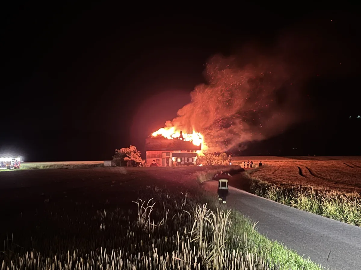 Nachts lodern Flammen aus dem Dach eines Gebäudes auf einem Feld. Rechts sind Feuerwehrleute im Einsatz.