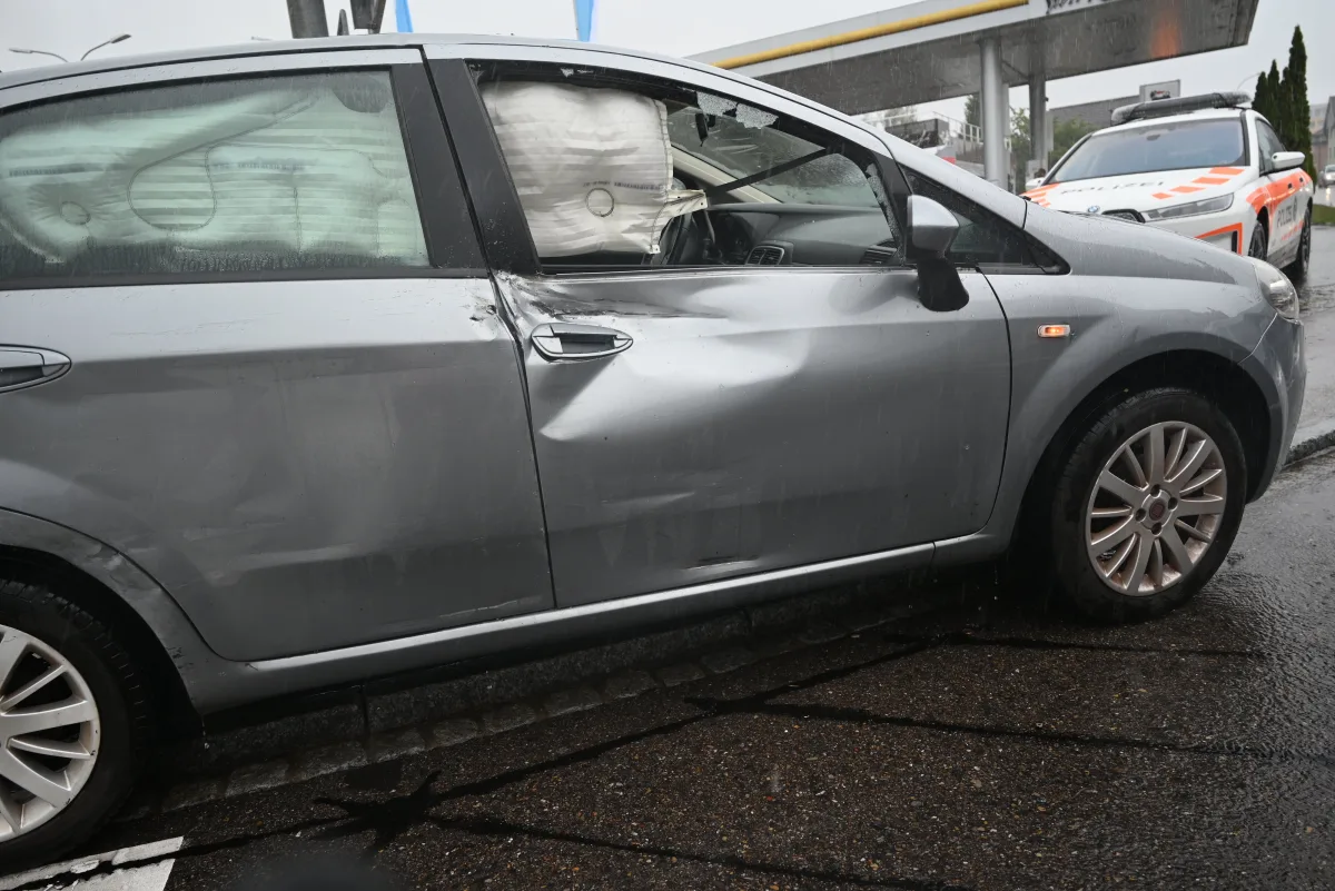 Ein silbernes Auto zeigt deutliche Unfallspuren an der Seite und hat einen geöffneten Seitenairbag. Im Hintergrund ist ein Polizeifahrzeug zu sehen.