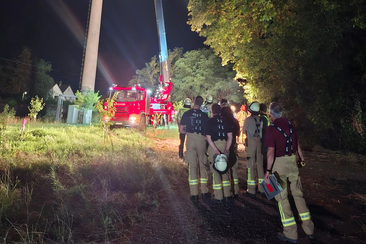 Eine Gruppe von Feuerwehrleuten steht bei Nacht in einer grasbewachsenen Umgebung. Im Hintergrund ist ein rotes Feuerwehrauto und ein Kran zu sehen, der an einem hohen Turm arbeitet.