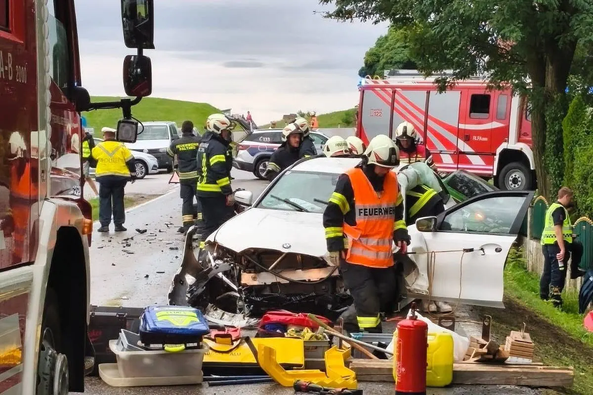 Ein schwer beschädigtes weißes Auto steht auf einer Landstraße, umgeben von Feuerwehrleuten und Polizeifahrzeugen. Rettungsgeräte sind am Boden verstreut.