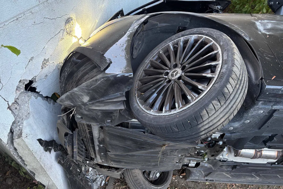 Ein schwarzes Auto ist nach einem Unfall seitlich gegen eine Wand gelehnt, wobei sichtbarer Schaden am Fahrzeug und an der Wand zu sehen ist.