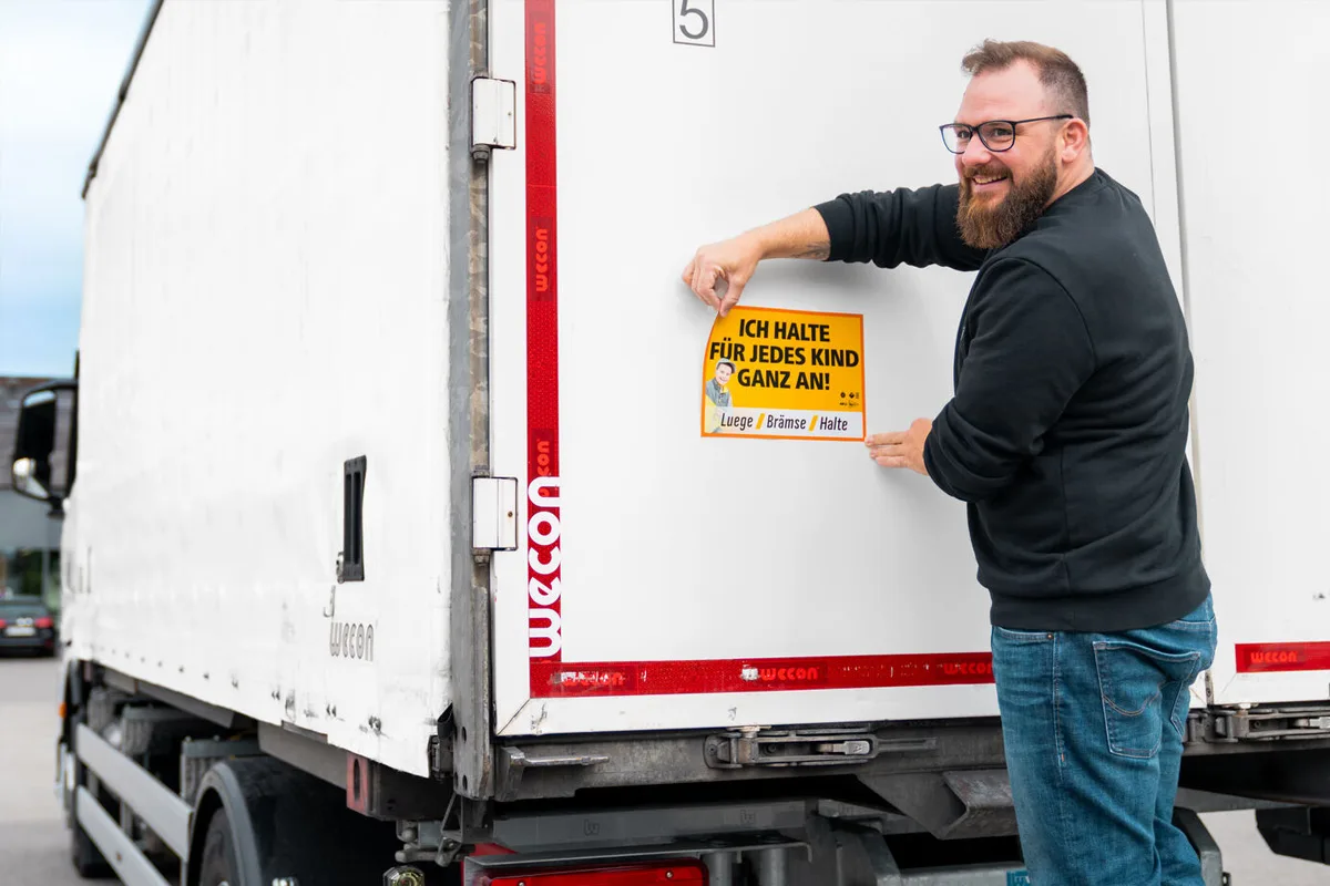 Ein Mann in dunkler Kleidung klebt ein gelbes Schild mit der Aufschrift 'Ich halte für jedes Kind ganz an!' auf einen weißen LKW.