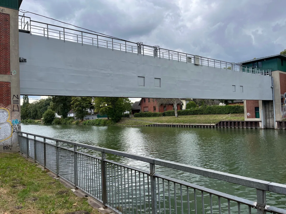 Eine graue Brücke überspannt einen ruhigen Fluss bei bewölktem Wetter. Am Ufer sind Bäume und Gebäude zu sehen.