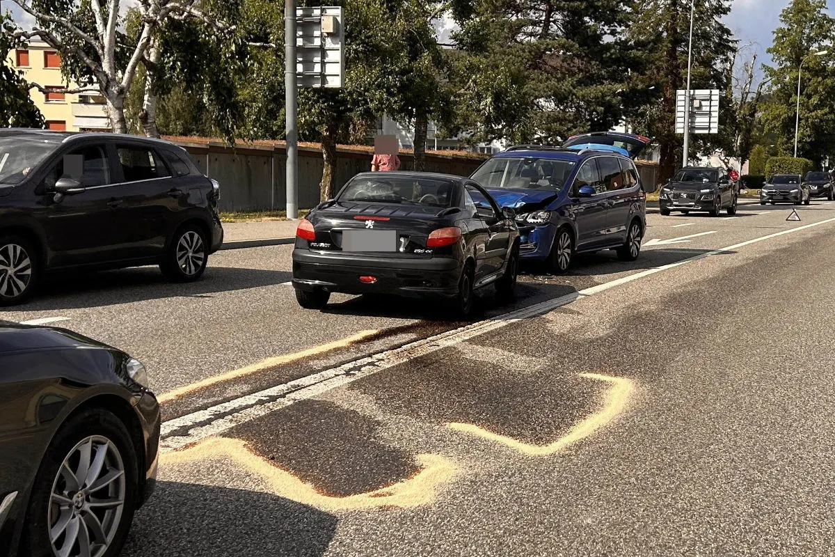 Ein blauer PKW mit sichtbarem Frontschaden ist auf ein schwarzes Fahrzeug auf einer Straße aufgefahren. Mehrere Fahrzeuge sind beteiligt, und die Straße ist mit Sand bedeckt, vermutlich zur Bindung ausgelaufener Flüssigkeiten.