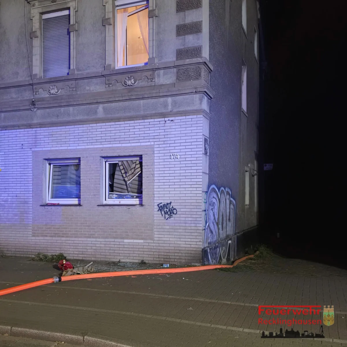 Ein Gebäude mit beschädigtem Fenster und einem Feuerwehrschlauch auf dem Gehweg. Das Gebäude hat Graffiti an der Wand.