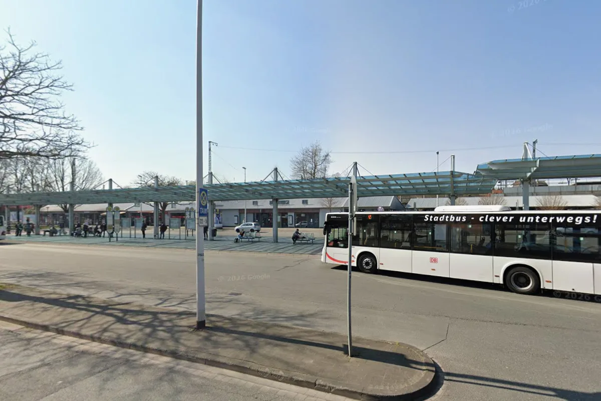 Ein Stadtbus steht an einer Haltestelle nahe eines Bahnhofs, umgeben von Menschen und Bäumen bei sonnigem Wetter.
