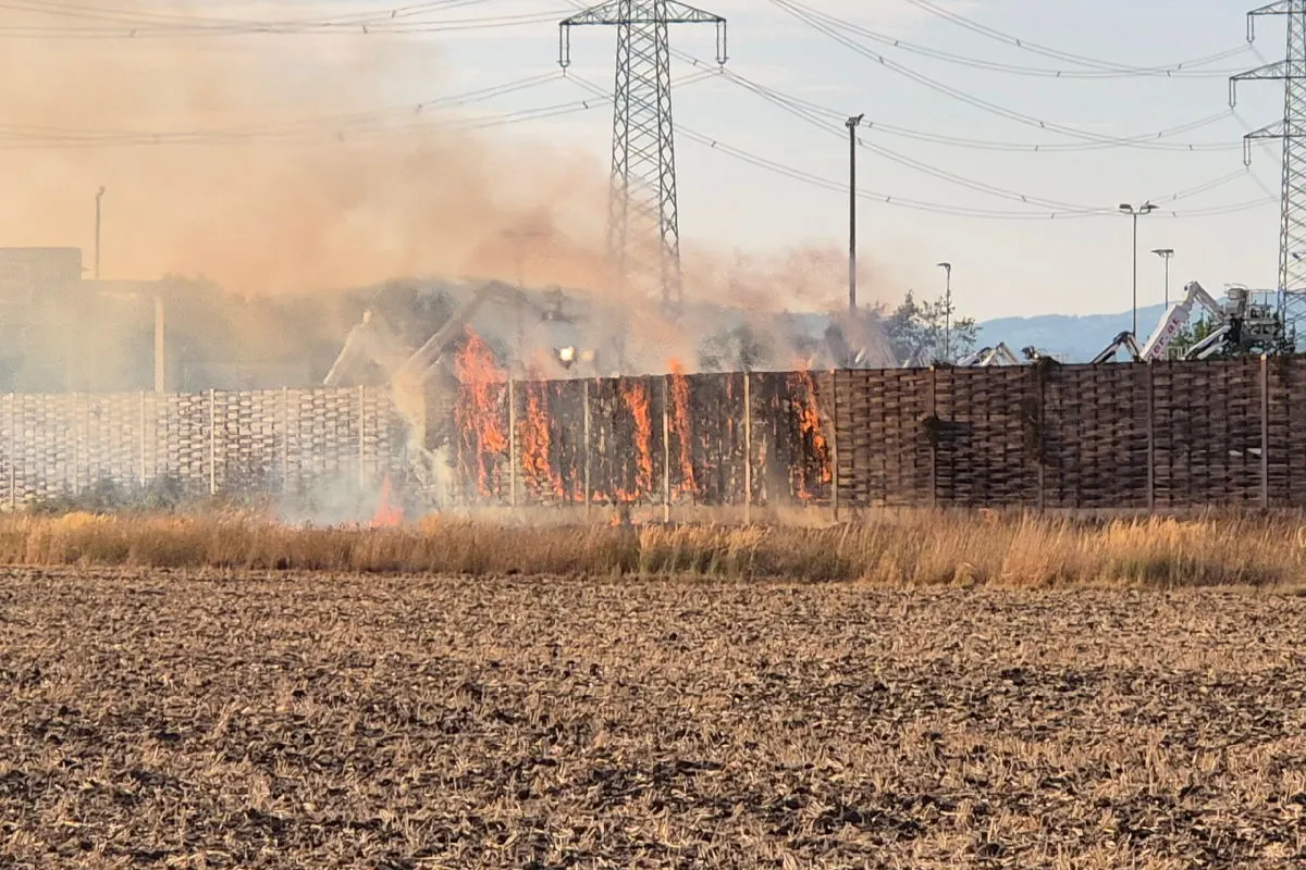 Ein großer Stapel Holzpaletten steht in Flammen auf einem Feld. Rauch steigt in den Himmel auf.