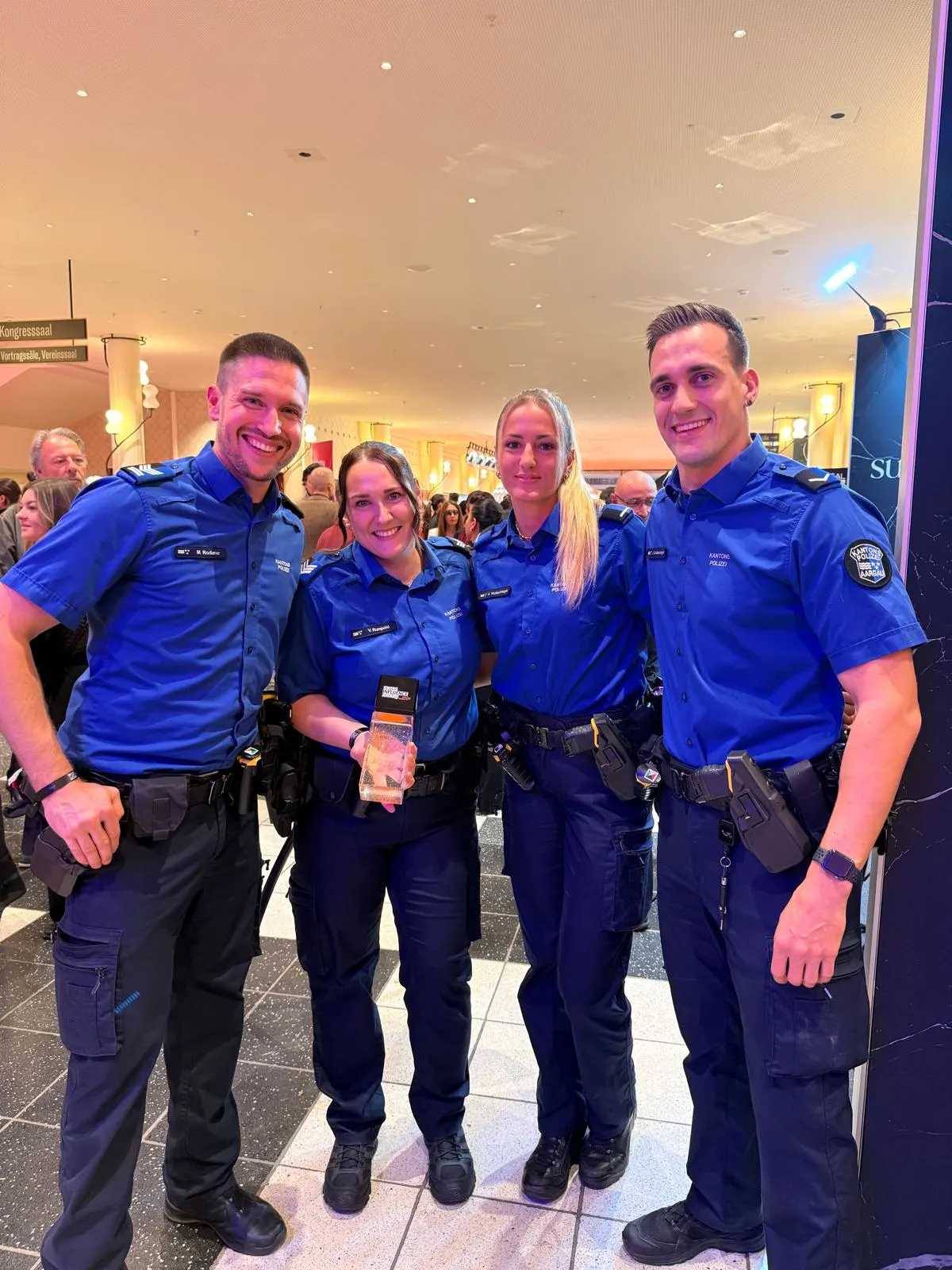 Vier Polizisten tragen blaue Uniformen und stehen lächelnd nebeneinander in einem hell erleuchteten Raum mit anderen Menschen im Hintergrund.