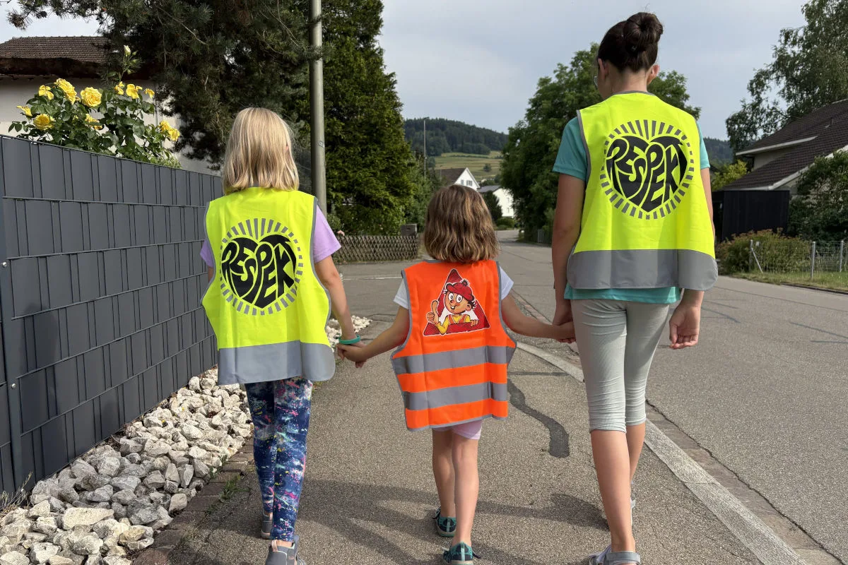 Drei Kinder in leuchtenden Sicherheitswesten gehen Hand in Hand auf einem Gehweg entlang einer ländlichen Straße.