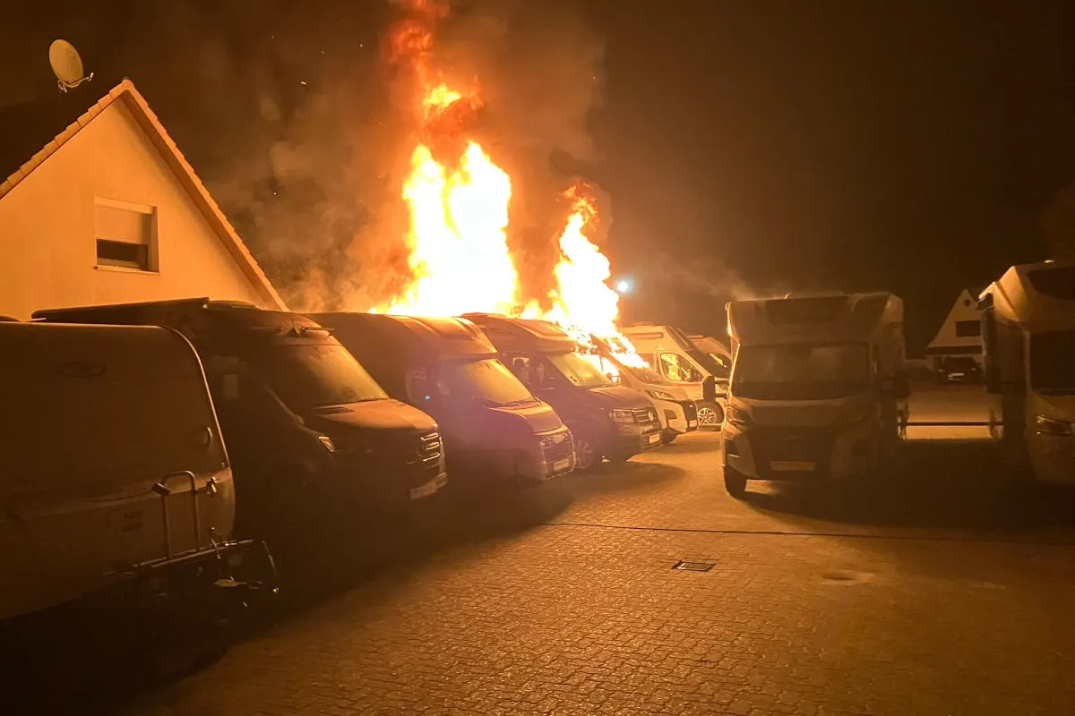 Mehrere Wohnmobile stehen auf einem Parkplatz, eines davon ist in große Flammen gehüllt. Es ist Nacht, und das Feuer erhellt die Umgebung.