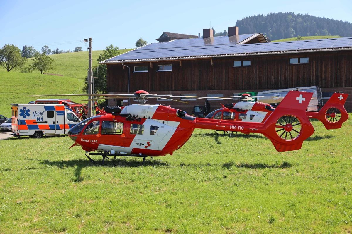 Auf einem grünen Feld stehen zwei Rega-Hubschrauber, während ein Rettungswagen im Hintergrund zu sehen ist. Die Szene wirkt ruhig und sonnig.
