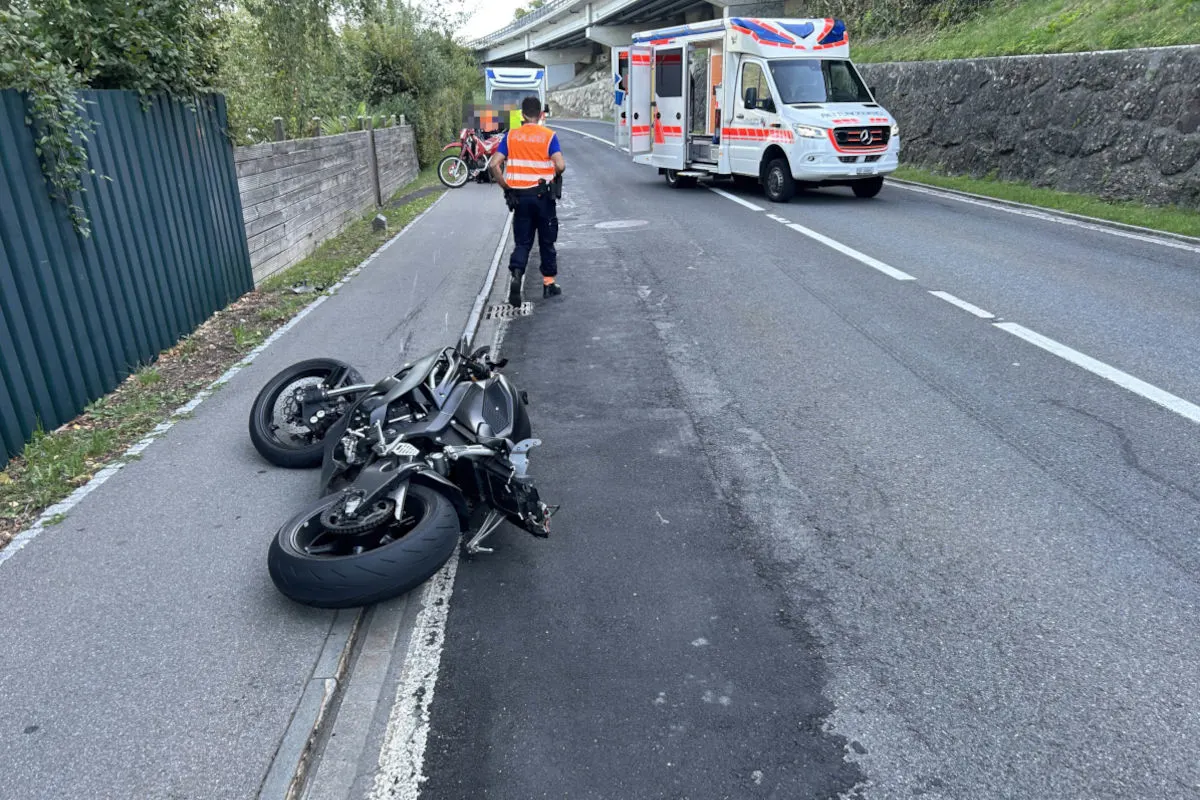 Ein schwarzes Motorrad liegt umgestürzt auf einem Gehweg neben einer Straße, ein Polizeifahrzeug und eine Ambulanz sind ebenfalls sichtbar.