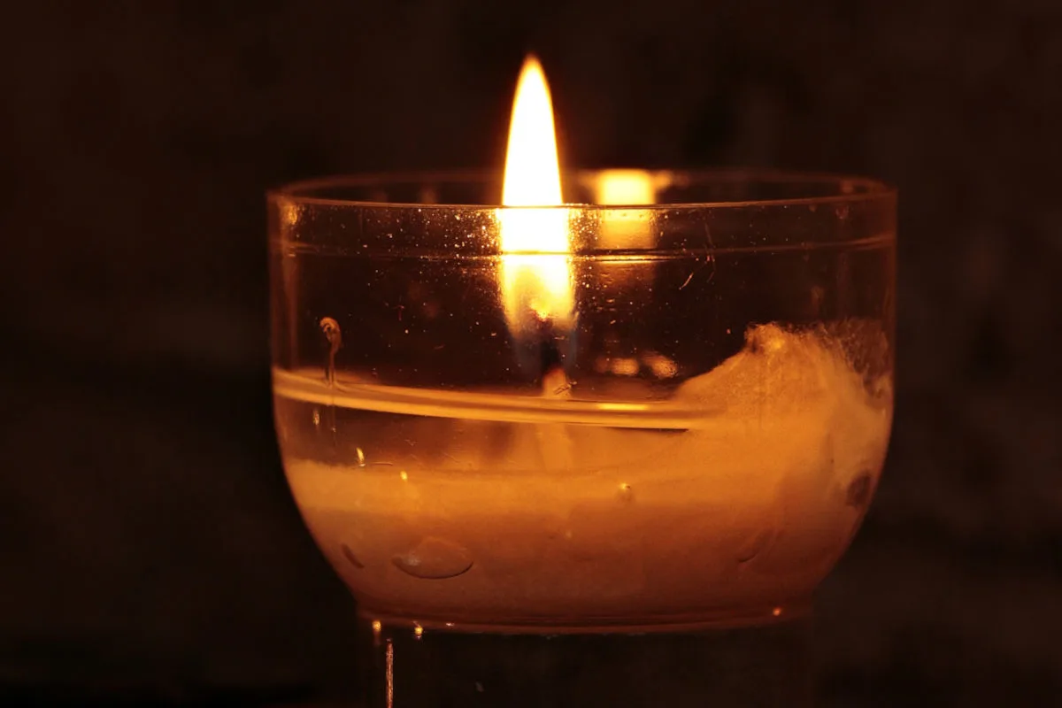 Eine einzelne Kerze brennt in einem durchsichtigen Glasbehälter, der ein warmes, gelbes Licht ausstrahlt. Die Umgebung ist dunkel, wodurch die Flamme hervorgehoben wird.