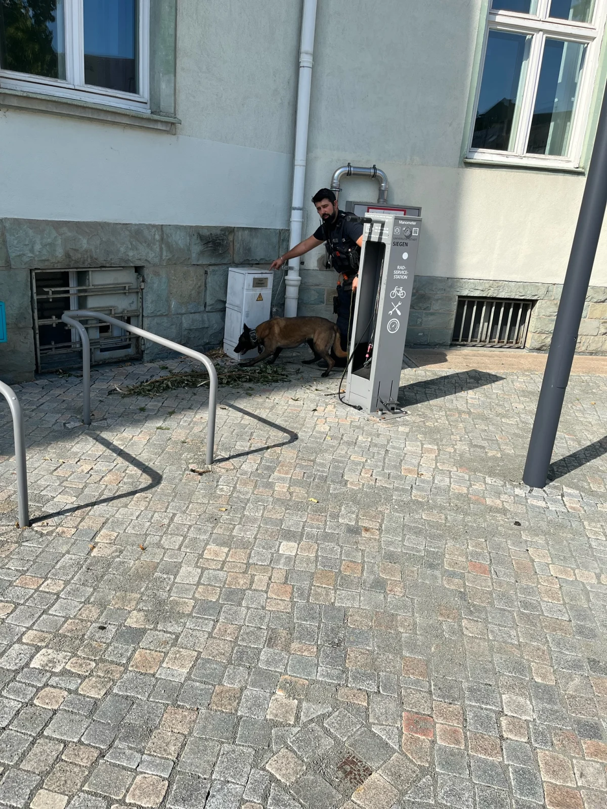 Ein uniformierter Polizist mit einem Diensthund steht neben einem Gebäude und inspiziert einen Bereich. Das Wetter ist klar.