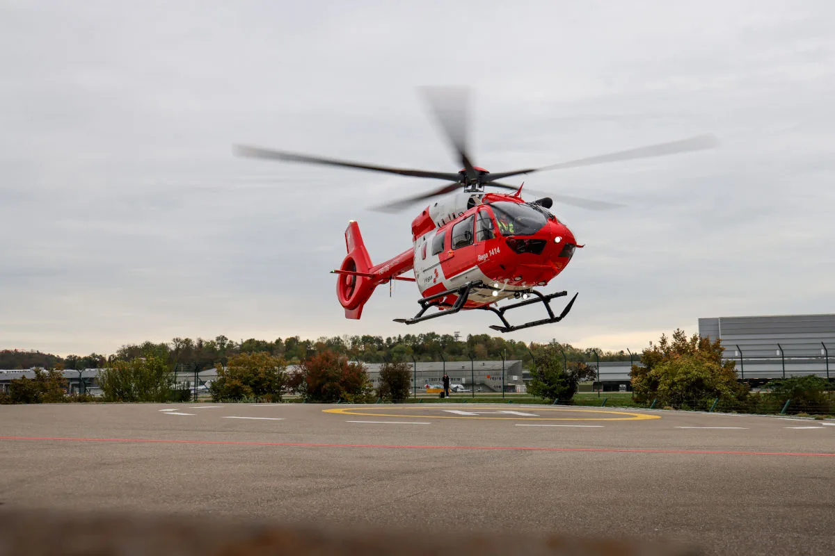Ein roter Rettungshubschrauber ist im Begriff abzuheben. Im Hintergrund ist ein bewölkter Himmel und Vegetation sichtbar.