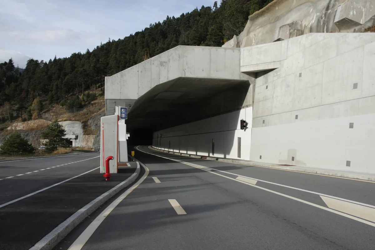 Eine leere Landstraße führt in einen Tunnel, umgeben von bewaldeten Hügeln. Der Himmel ist teilweise bewölkt.