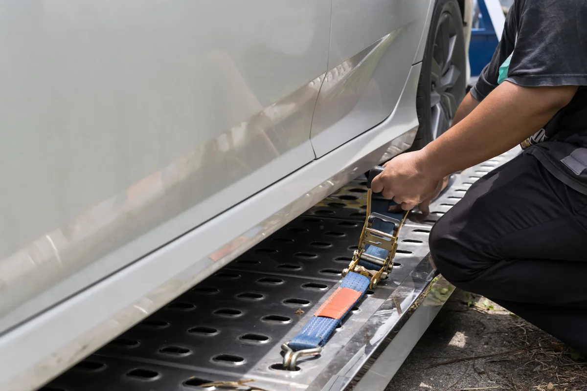 Eine Person befestigt einen blauen Spanngurt an einem weißen Auto auf einem Abschleppwagen. Das Fahrzeug steht auf einer metallenen Plattform.