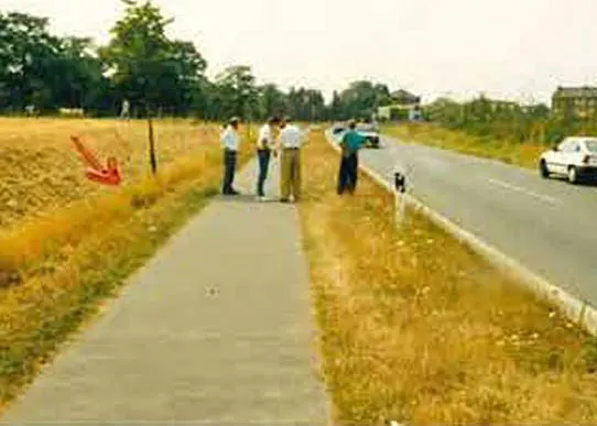 Mehrere Personen stehen an einer Landstraße, während ein Auto die Straße entlangfährt.