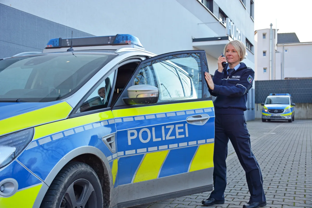 Eine Polizistin steht neben einem blau-gelben Streifenwagen vor einem Polizeigebäude und spricht in ein Funkgerät. Im Hintergrund ist ein weiteres Polizeifahrzeug sichtbar.