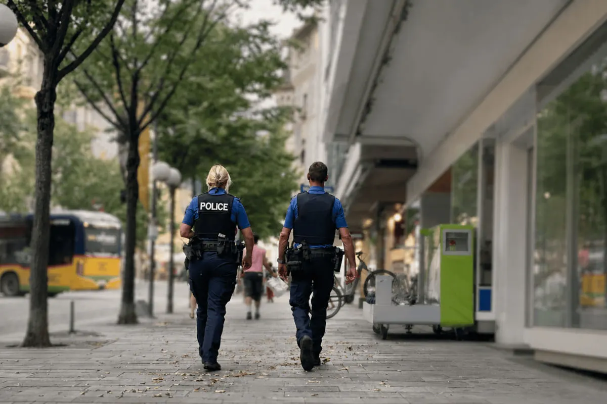 Zwei Polizisten in blauer Uniform mit Westen und Ausrüstung gehen auf einem Gehweg in einer städtischen Umgebung, flankiert von Bäumen und Geschäften.