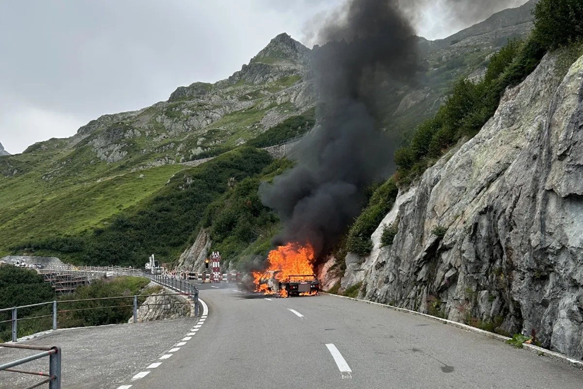 Ein Auto steht in Flammen auf einer kurvenreichen Bergstraße, umgeben von Felsen und Vegetation, während schwarzer Rauch aufsteigt.