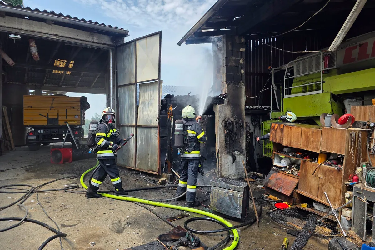Zwei Feuerwehrleute in Schutzkleidung löschen mit Schläuchen einen Brand in einer Werkstatt. Rauch ist sichtbar.